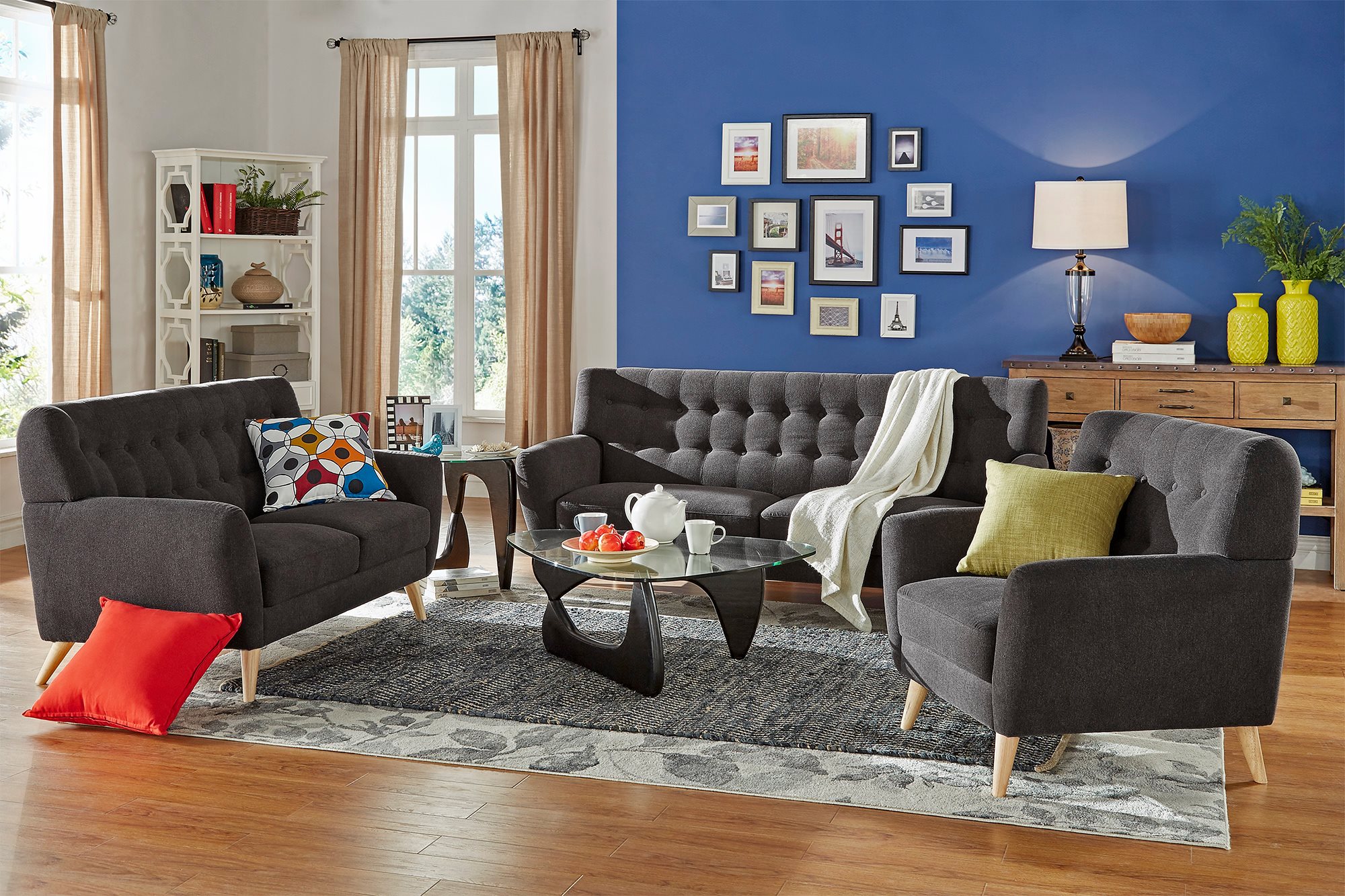 This is the last image for home design trends. It displays a mid-century modern themed living room. There is a sofa, loveseat, and accent chair with dark grey linen upholstery and natural finish peg feet. There are glass tables and colorful accent pillows.