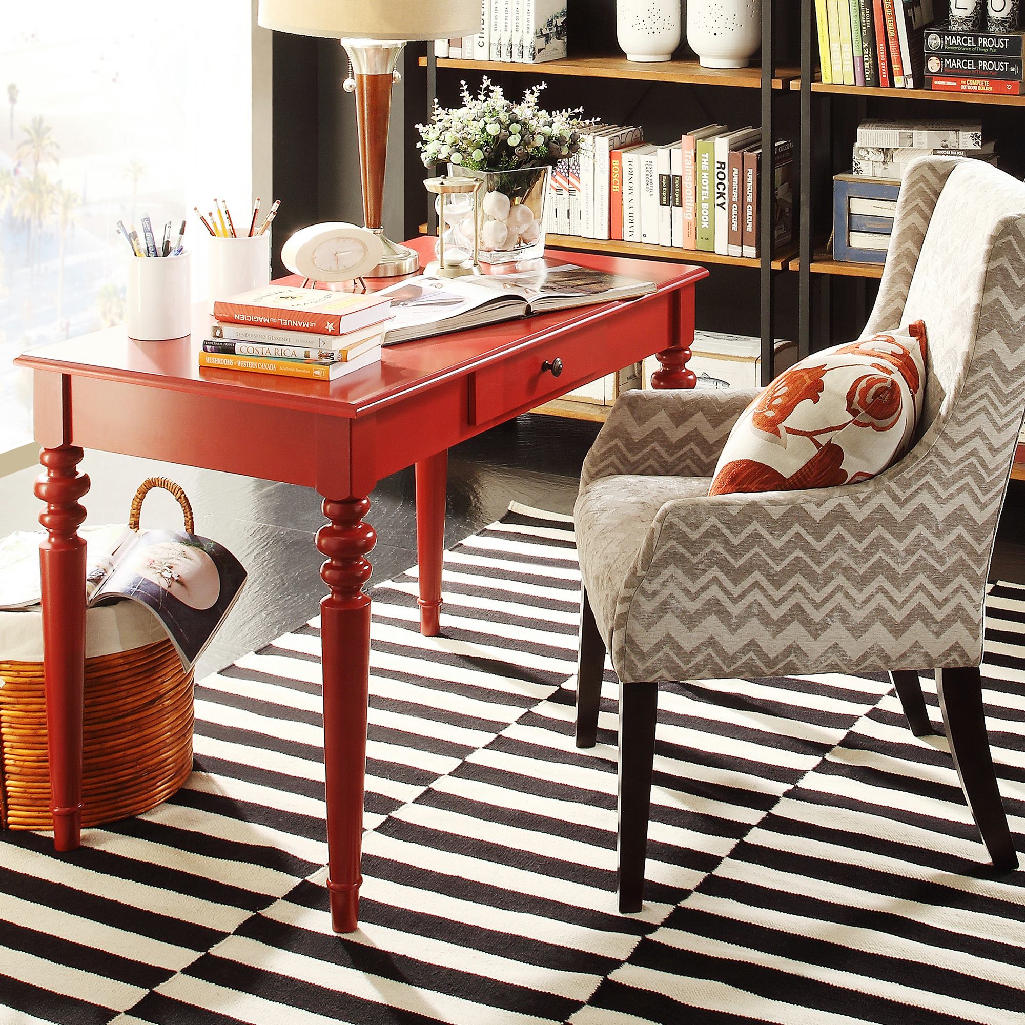 Home décor trends are all about having fun, and this image depicts a mixture of patterns. There is a black and white striped rug, a grey chevron printed hostess chair, and a bright red desk. The chair also has a red floral-printed accent pillow on it.