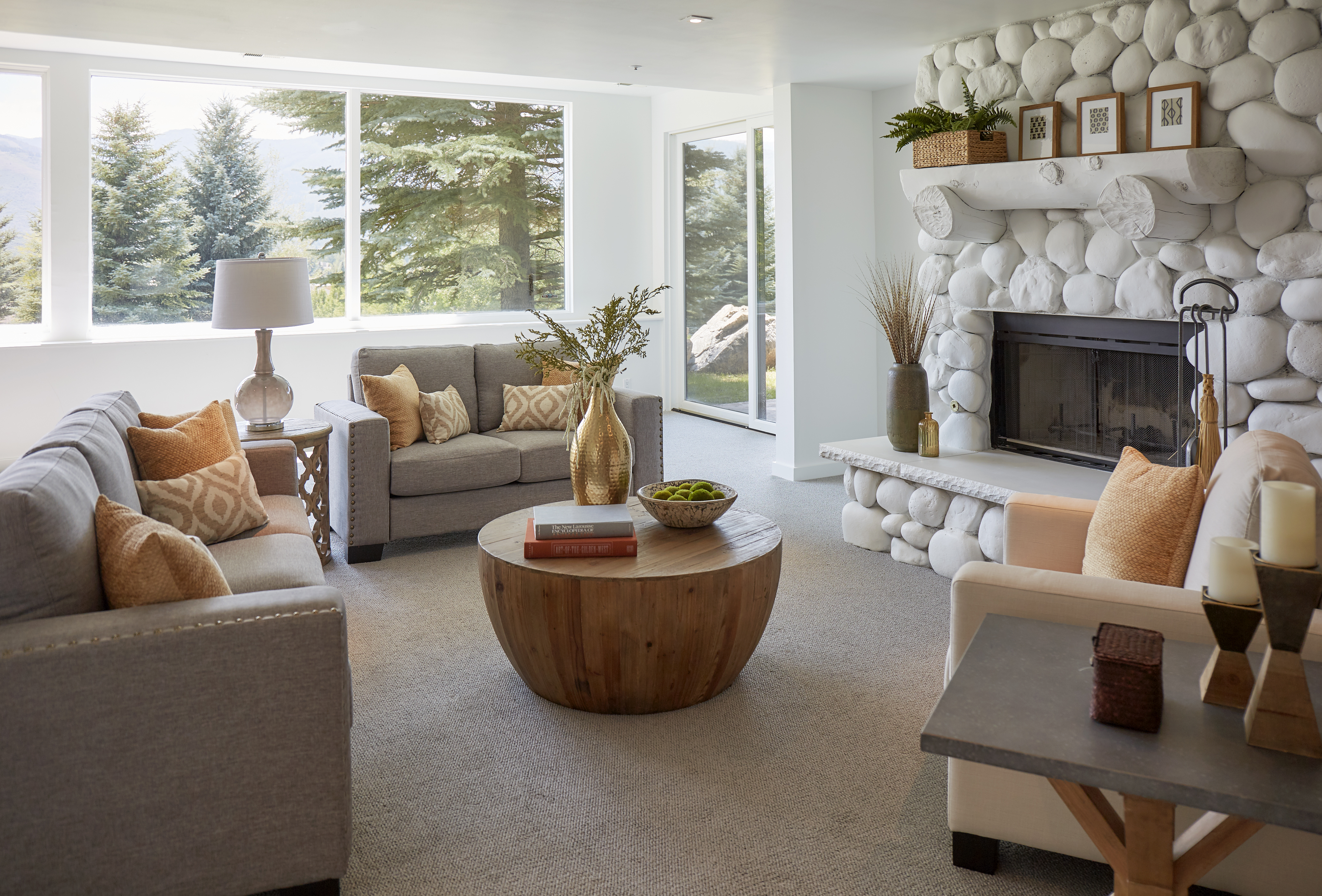 This modern and rustic space is a living room. There is a white stone and wood fireplace off to the side. There are grey linen upholstered sofa and loveseat and a white linen upholstered accent chair. The coffee table is a reclaimed wood drum-shaped coffee table. 