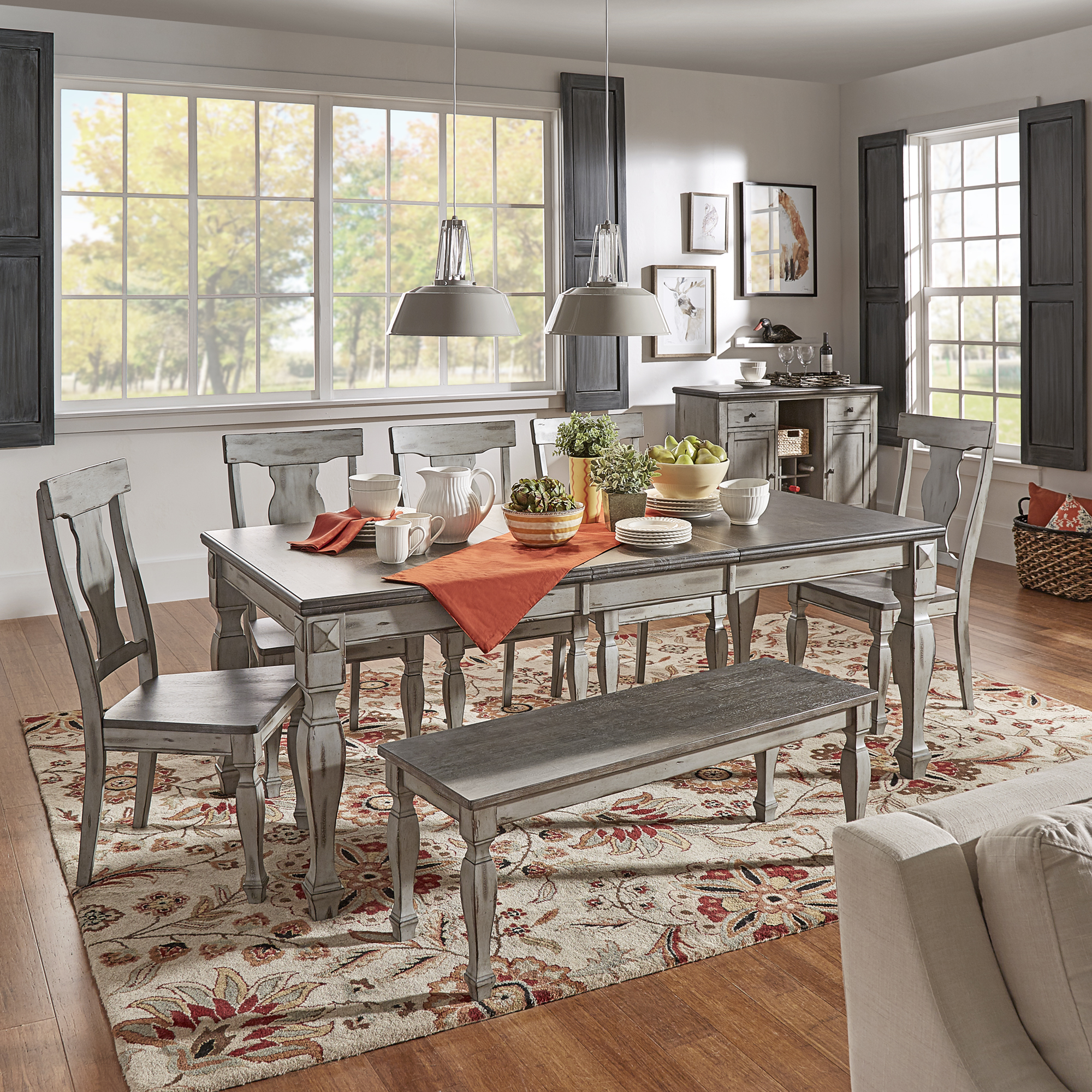 This image shows the iNSPIRE Q Grey Two-Tone Antique Finish Wood Dining Set with 5 chairs, 1 bench, and 1 dining table. The dining table is decorated with white, yellow, green, and orange accessories. The white and orange accessories complement the red and white rug on the hardwood floors.