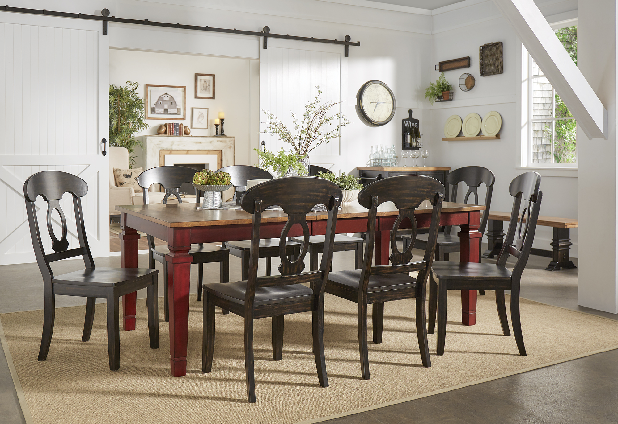 This one of the 10 inspiring dining combinations puts a modern twist on a timeless classic. The center of the room has a long, rectangular dining table with an antique berry red finish base and oak finish table top. Placed around the dining table are 8 dining chairs in a dark brown finish, standing out against the colors of the dining table and the white barndoors in the background.