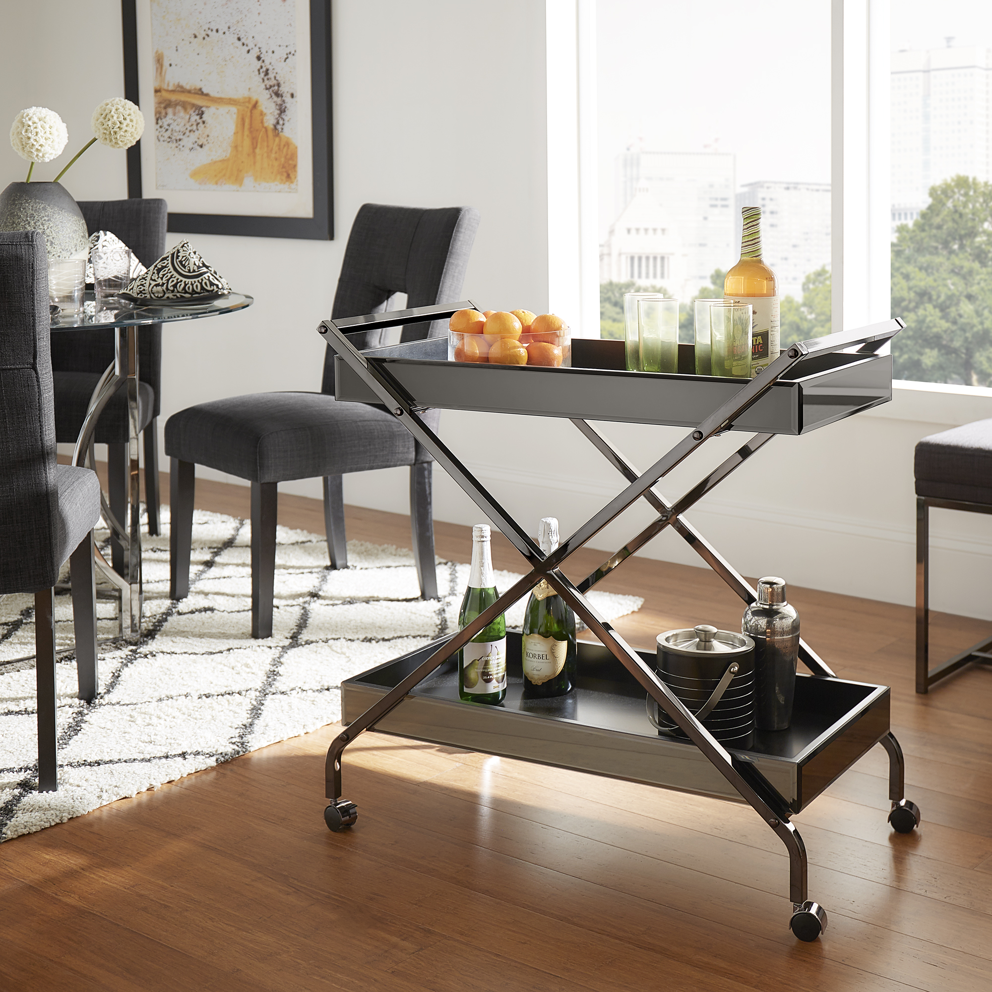 In this image, we use the same bar cart to display a different way as to how to style a bar cart. This bar cart still has the mirror sides and chrome frame. The décor has a lot more color, as the top shelf has a bowl of oranges, green glasses, and an orange bottle. The bottle shelf has green bottles and metal barware.