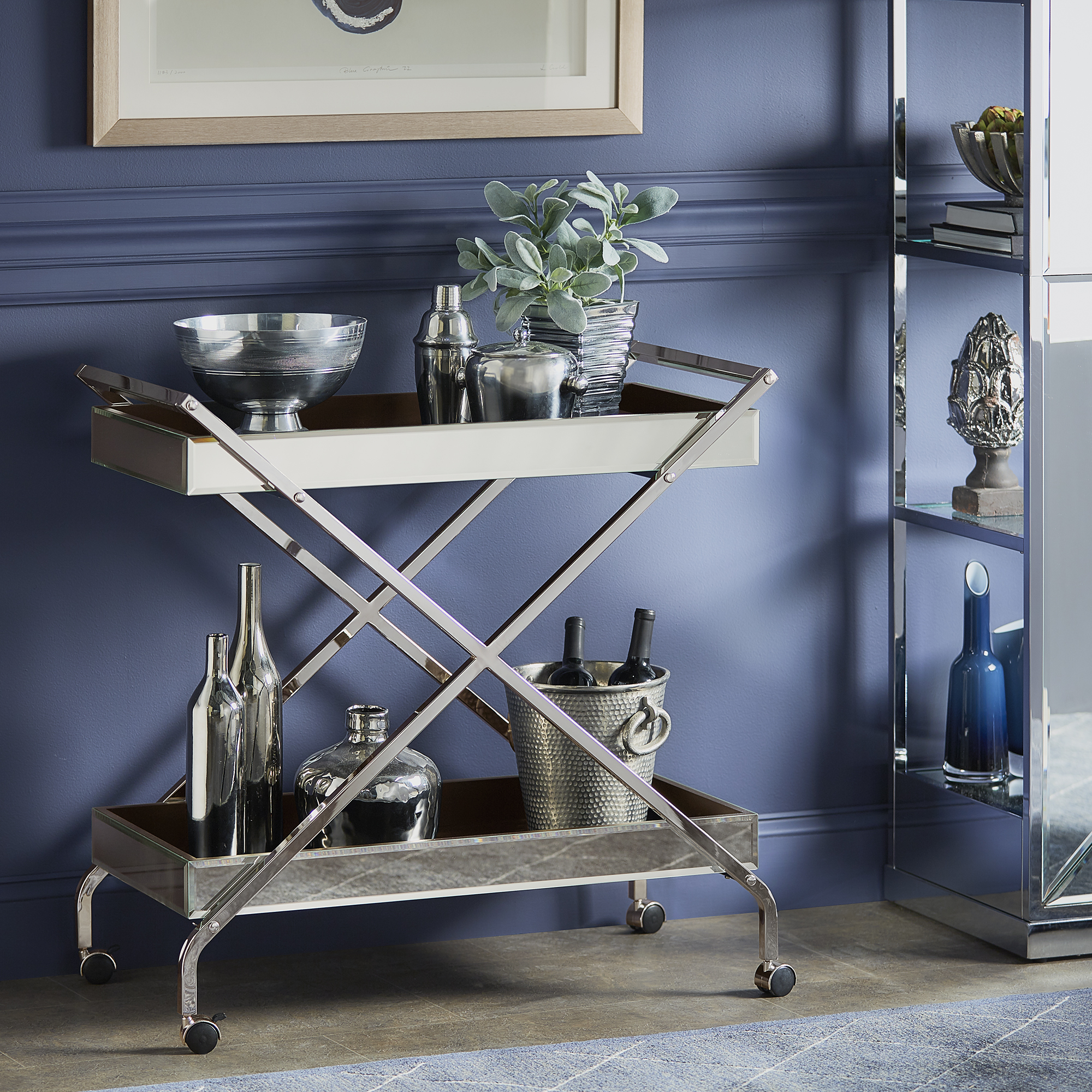 This first image used to depict how to style a bar cart features a glam-inspired bar cart. The bar cart has shiny chrome finishes and is decorated with chrome and silver barware. A potted plant on the top shelf adds a pop of color.