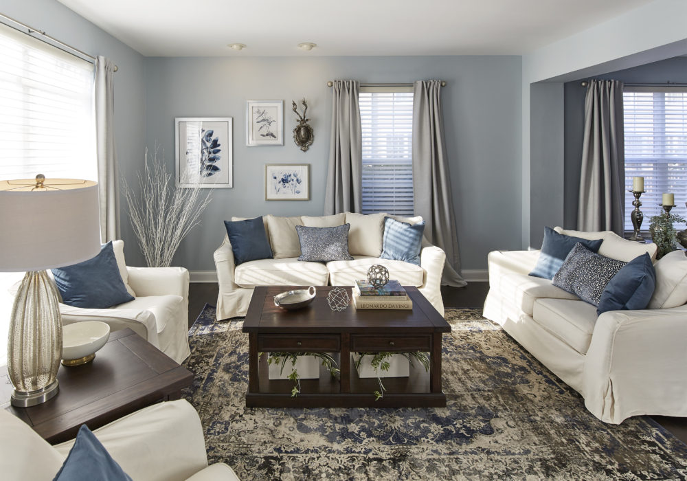 This image focuses solely on the formal living room. There is plenty of plush, white seating with blue accent pillows. A rich brown coffee table and matching end table add a touch of warmth and ties in with the browns of the rug.