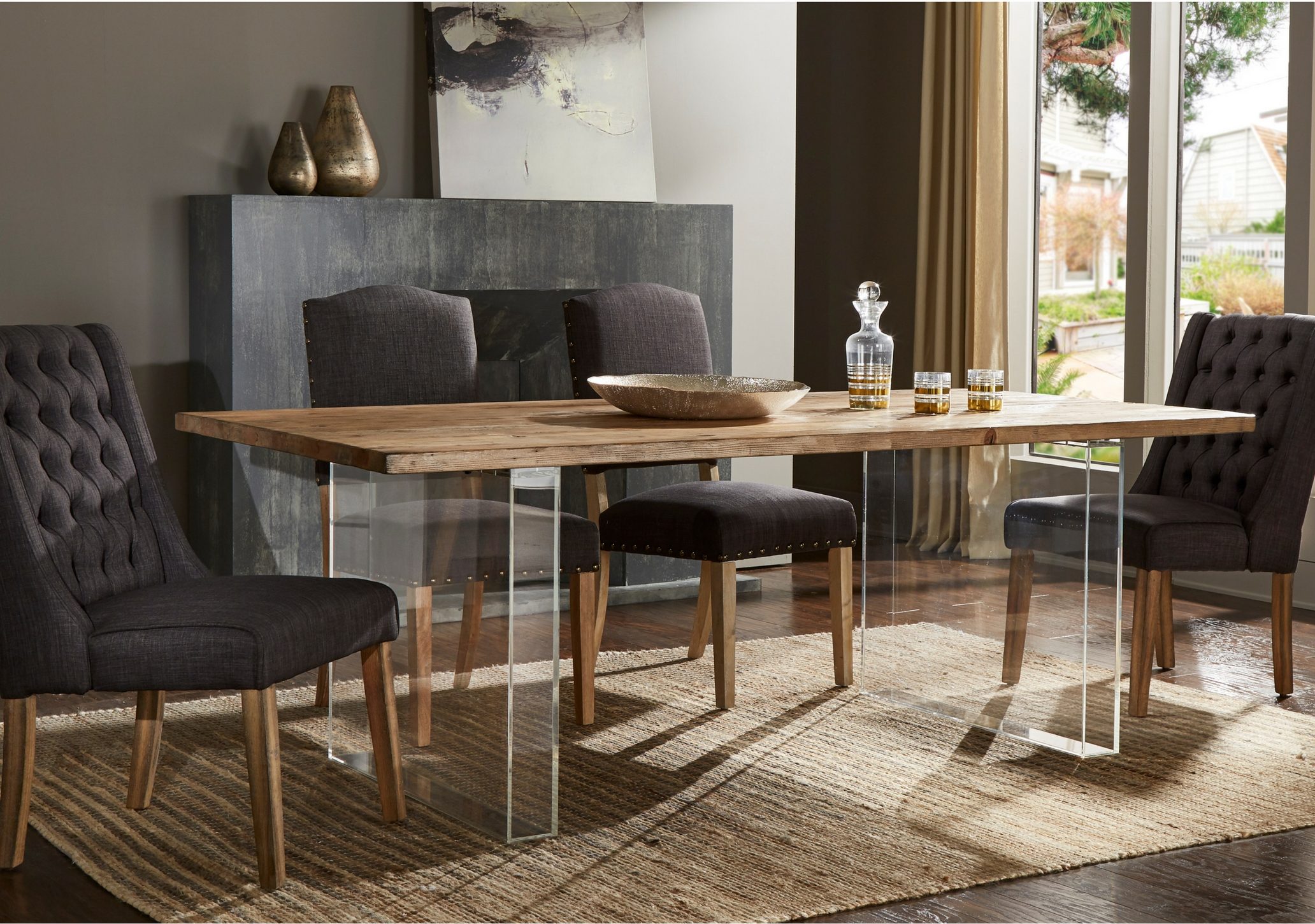 Blending artisan with modern, this one of the 10 inspiring dining combinations features a dining table with a reclaimed wood table top and clear acrylic block legs. Surrounding the table are four dark grey linen upholstered chairs. The table top is decorated minimally with a few glasses and a decanter.