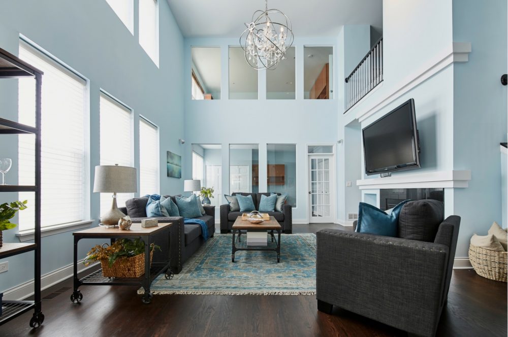 The full view of the great room. Like in the front of the house, the walls are light blue. The sofa, loveseat, and accent chair are upholstered in dark grey linen and paired with blue accent pillows. The tables and bookcase in the room are made with wood and metal for mixed media appeal.