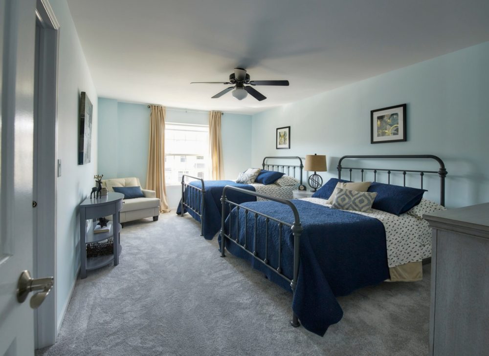 The guest bedroom features two farmhouse-themed metal full beds. The bedding is dark blue and white. Other pieces include grey wood nightstand, sofa table, and dresser.