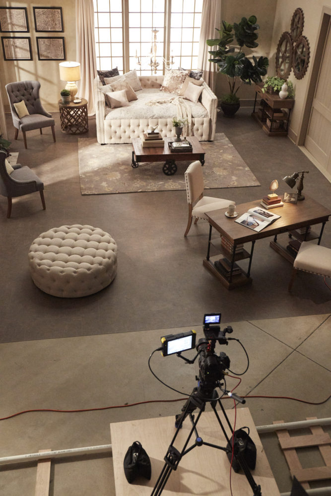This image shows a more aerial view of our formal living room setup for filming. At the bottom of the image is the video camera, prepped for recording. The rest of the image is the living room, including the beige linen tufted round cocktail ottoman, grey linen tufted wingback chairs, beige linen tufted daybed, wood and metal coffee table, beige linen nailhead side chairs, and cornice 1-drawer writing desk.