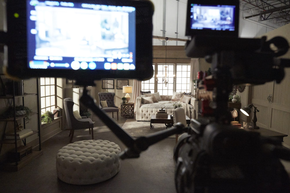This image shows our formal living room setup through the lens of a video camera. In the foreground is the video camera, turned on and ready to start filming. In the background, we see the iQ artisan pieces, including a beige linen round tufted ottoman, grey linen tufted wingback side chairs, beige linen tufted daybed, and a wood and metal coffee table.