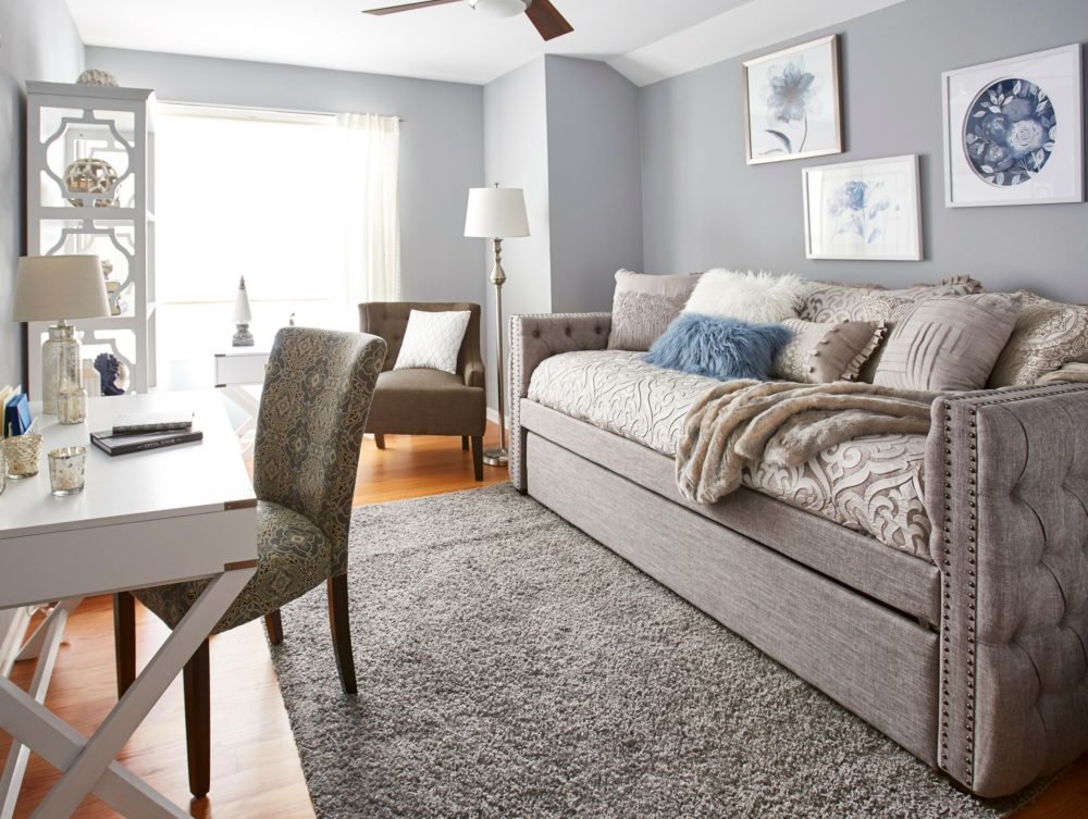 The full view of the spare room. To the left is the white desk, printed desk chair, and white bookcase. On the right wall is a grey fabric weave daybed with trundle. The daybed has button tufting and nailhead trims for added glam detail. It is dressed with grey and beige begging and pillows. In the back of the room is a sloped arm accent chair and a white campaign end table.