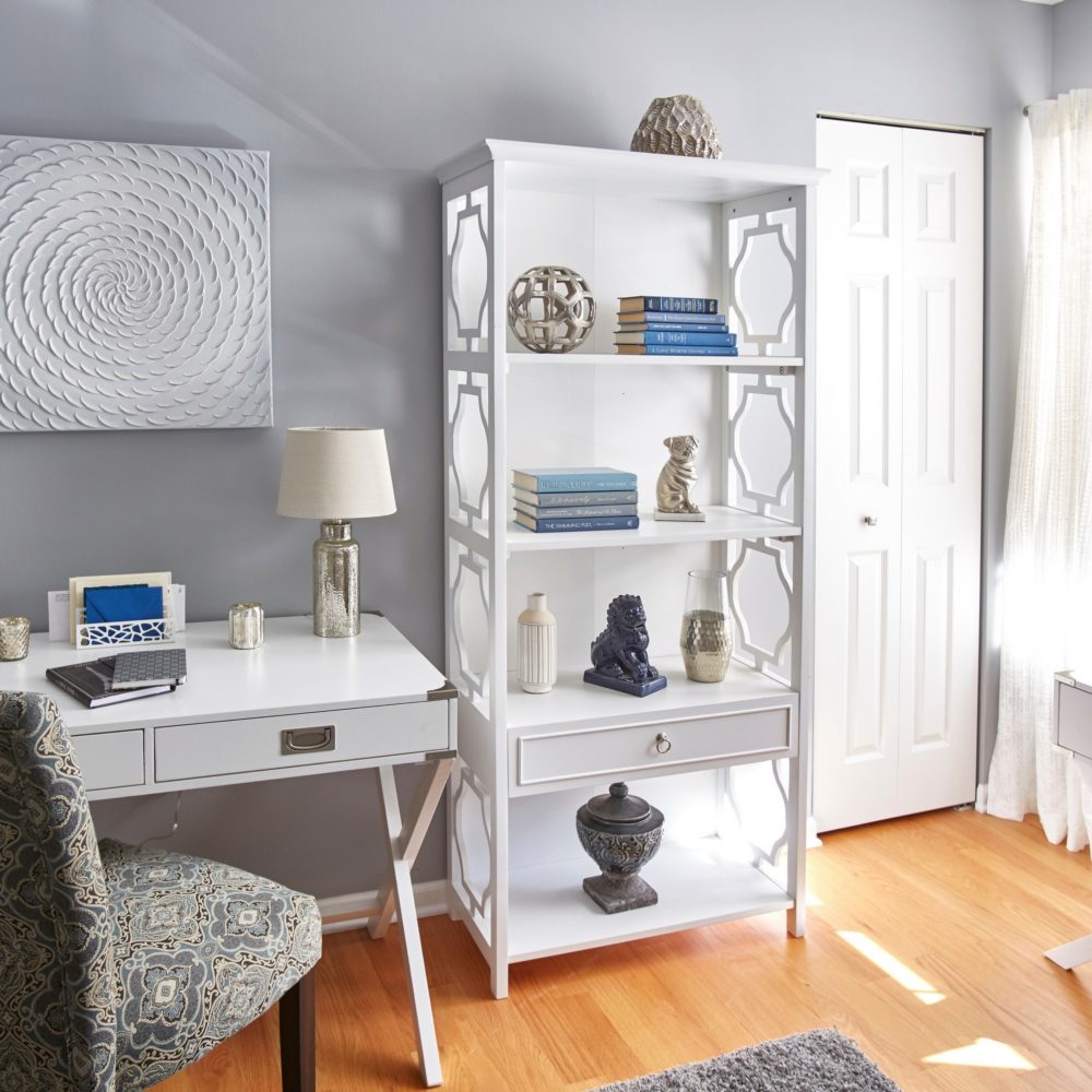This image shows one wall of the apre room. There is a white-finished campaign writing desk. There is a printed upholstered parsons chair in front of the desk. Beside the desk is a 4-shelf white bookcase, complete with one drawer. This bookcase features blue, white, and grey books and décor. 
