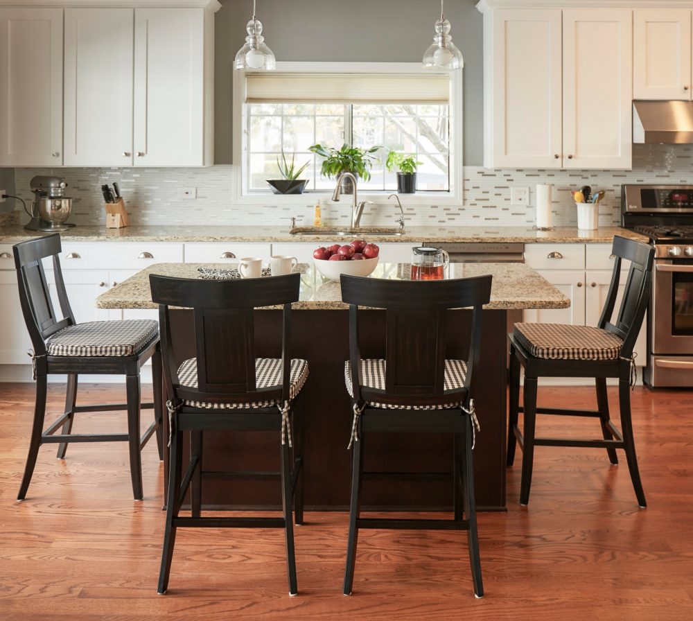 This is the kitchen in our Illinois home makeover. There is an island in the middle of the image, surrounded by four counter height stools. The stools are wood, with panel backs, antique black finish, and black and white checkered patterned seat cushions. 