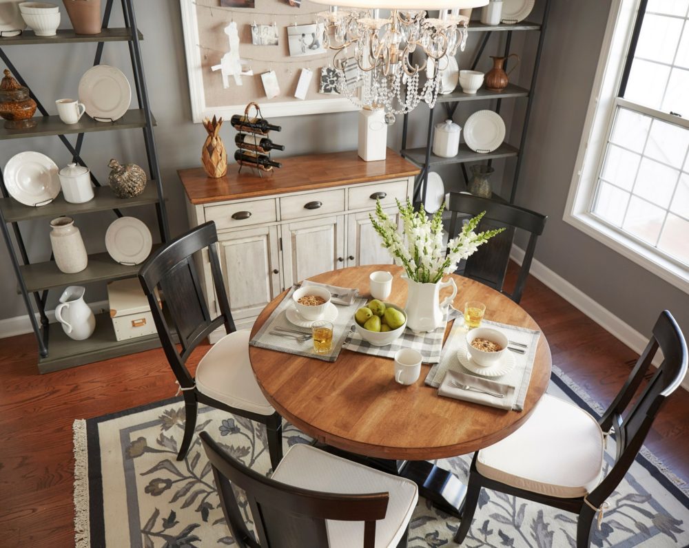 This a higher view of the farmhouse-themed eat-in kitchen. In the center is a round, oak-finished top dining table. It is decorated with white and grey place settings, as well as a vase of greenery. There are four wood dining chairs around the table, all in an antique black finish and with panel backs. Each chair has a plain white seat cushion. Behind the dining set is a two-tone antique finish buffet server. The top is oak, like the dining table, and the base is in antique white. On each side of the server is a grey-finished wood and metal bookcase displaying white china.