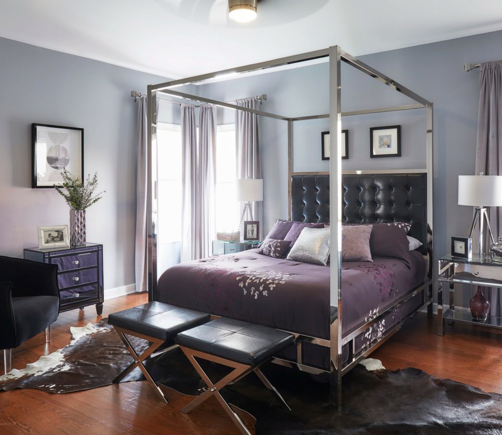 This is a wider view of the bedroom. We still see the canopy bed, the two footstools, and the three mirrored end tables. We also see an accent chair with acrylic legs and black velvet upholstery. On the floor is a faux cowhide print rug.