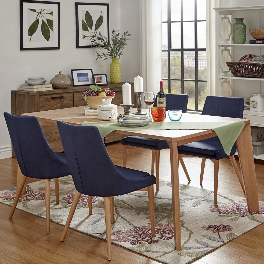 The last (but not least!) of our modern tablescape ideas. This is a wood, natural-finished dining table with four blue barrel back upholstered dining chairs. A green table runner is draped over one corner of the rectangular dining table. There is a variety of colorful dinnerware on the table, mainly white, blue, red, and orange.