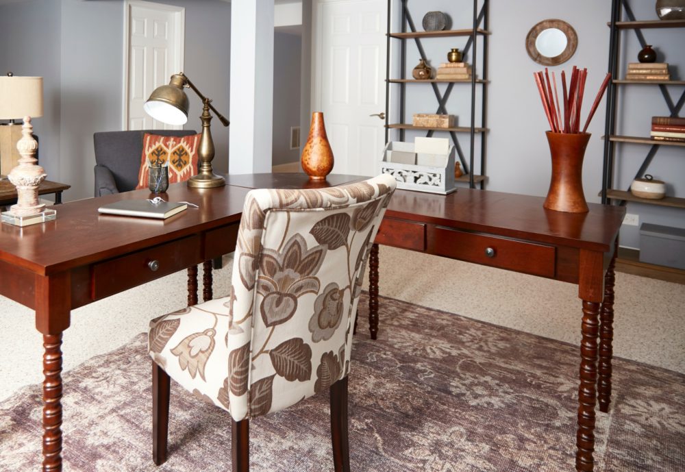 This views the home office from the opposite corner. In the center is the cherry-finished L-shaped corner desk with helix legs. In front of the desk is an upholstered parsons chair with a brown leaf print. The desk has an antique brass finished desk lamp and some brown pieces of décor. Behind the desk is a grey linen armchair. Opposite to the armchair are two wood and metal bookcases, decorated with books and pieces of décor. 