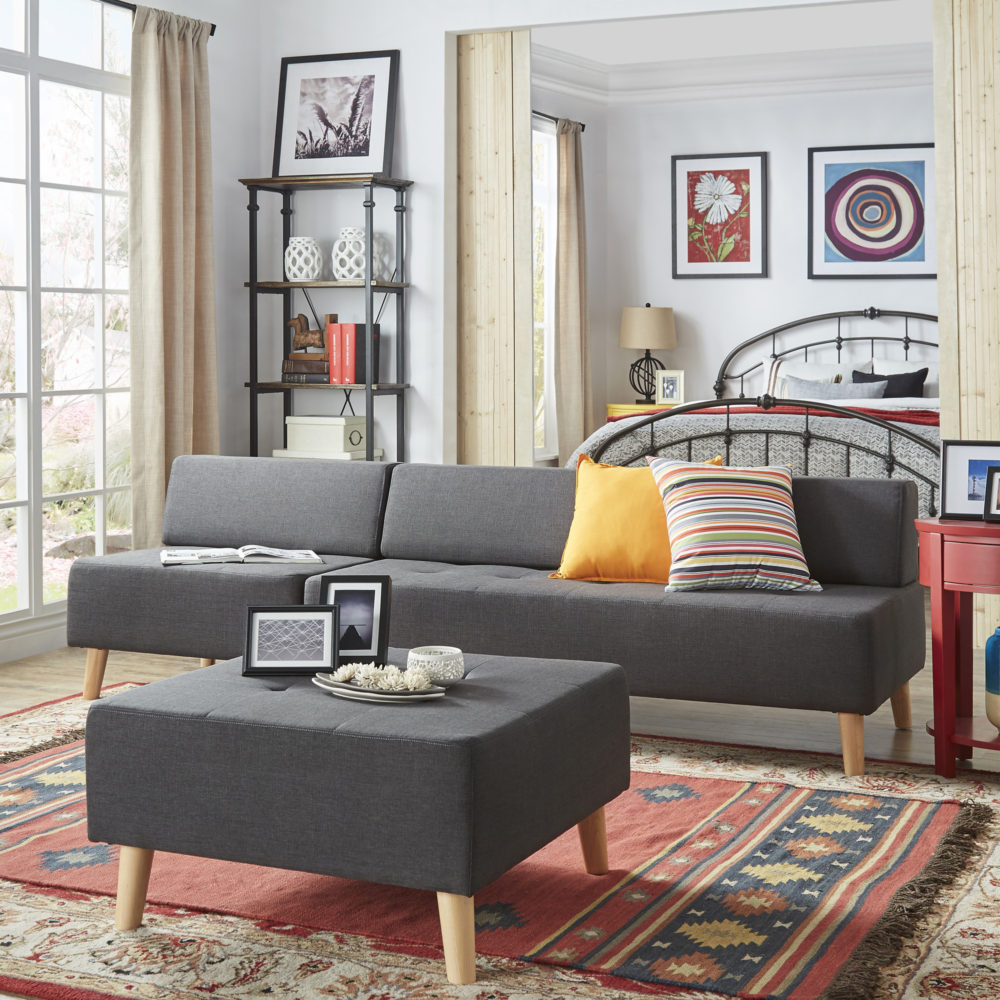 This living room features a grey linen upholstered armless sofa. It has two accent pillows, one yellow and one rainbow striped one. In front of the sofa is a matching grey linen square ottoman. Behind the sofa is a doorway to a bedroom with a metal bed. There is also a samba red end table.