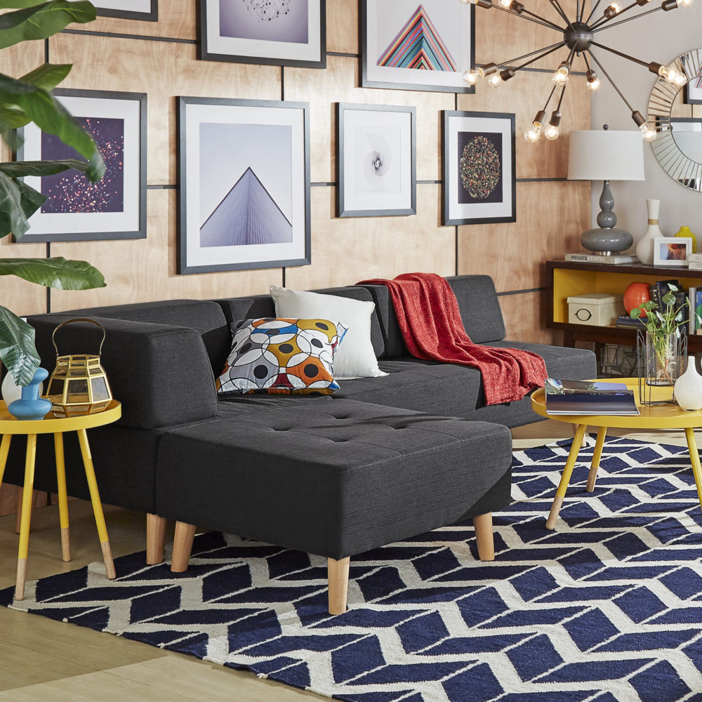 This mid-century modern living room has a dark grey linen armless sectional sofa with a chaise ottoman. Two white and printed accent pillows sit on the sofa, along with a red throw blanket. Bright yellow matching end table and coffee table add another pop of color to the overall space.