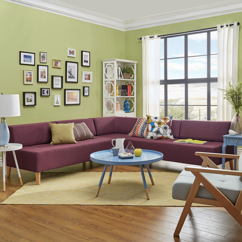 This colorful living room has green walls, a red sofa, a blue coffee table, a white side table, a blue end table, and a light blue accent chair. The tawny port red armless sectional sofa is in the heart of the room. Various, colorful pillows decorate the sofa, along with an open book.