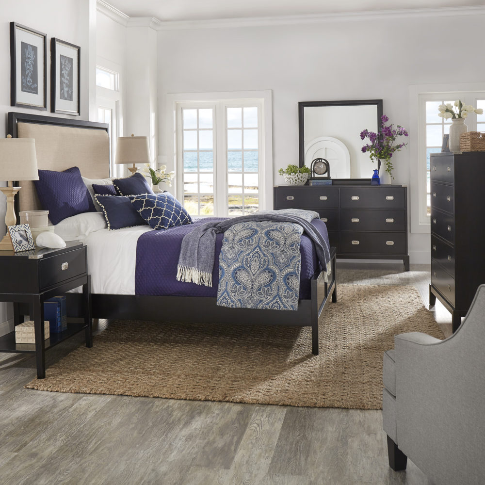 Now we display classic interior design styles for the bedroom. This room uses a fairly neutral color palette of black, white, and grey, with pops of color of blue. A black bed in the center of the room is dressed with blue, indigo, and white bedding. The coordinating nightstand, dresser, and chest, all come in a black finish with silver hardware. 