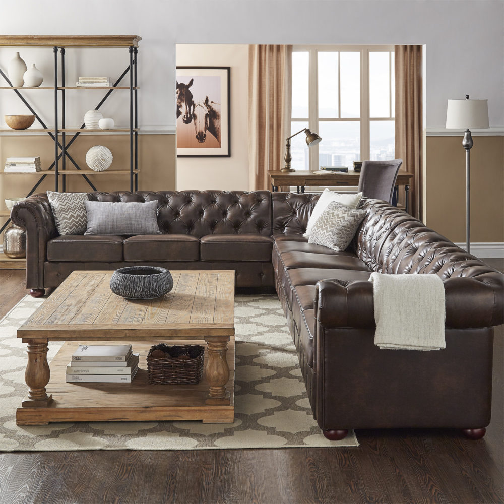This is a different look yet still offers the same refinement and Old World grandeur. The L-shaped chesterfield sectional sofa is upholstered in brown bonded leather with deep button tufting. The rectangular wood coffee table with baluster legs has a distressed brown finish that matches the brown finish of the wood and metal bookcase and desk in the background.