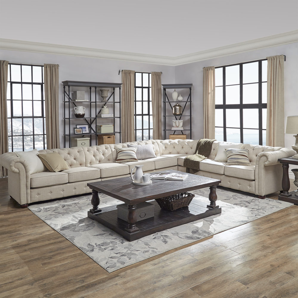 This living room setup features another L-shaped chesterfield sectional sofa with beige linen, button-tufted upholstery. The dark brown wood rectangular coffee table and matching end table offer a striking contrast against the lightness of the sofa. Behind the sofa are two wood and metal cornice bookcases with a grey wood finish.