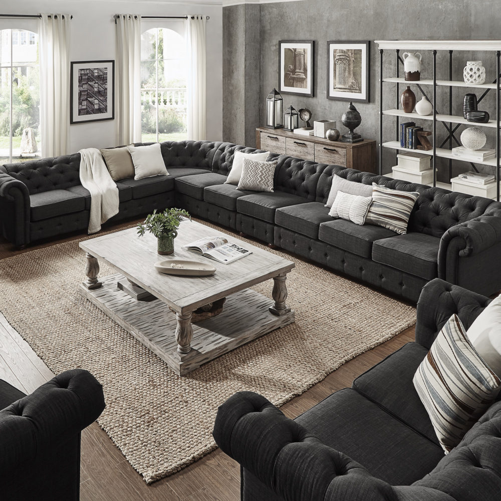 A large, living room setup with an L-shaped chesterfield sectional sofa. It is upholstered in dark grey linen and features plush button tufting. A rectangular wood coffee table with a white distressed finish and baluster-styled legs, further enhancing the look of Old World grandeur.