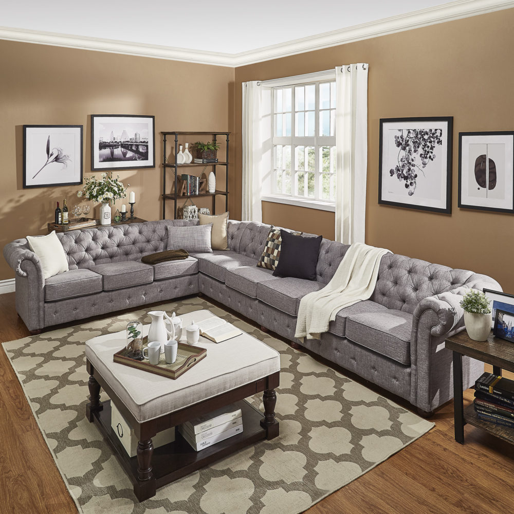 This last living room setup once again uses an L-shaped chesterfield sectional sofa with grey linen, button-tufted upholstery. In front of the sofa is a cocktail ottoman, with a rich dark brown wood base and light grey linen cushioned top. Rustic tones come from the wood and metal end table, bookcase, and sofa table surrounding the sofa.