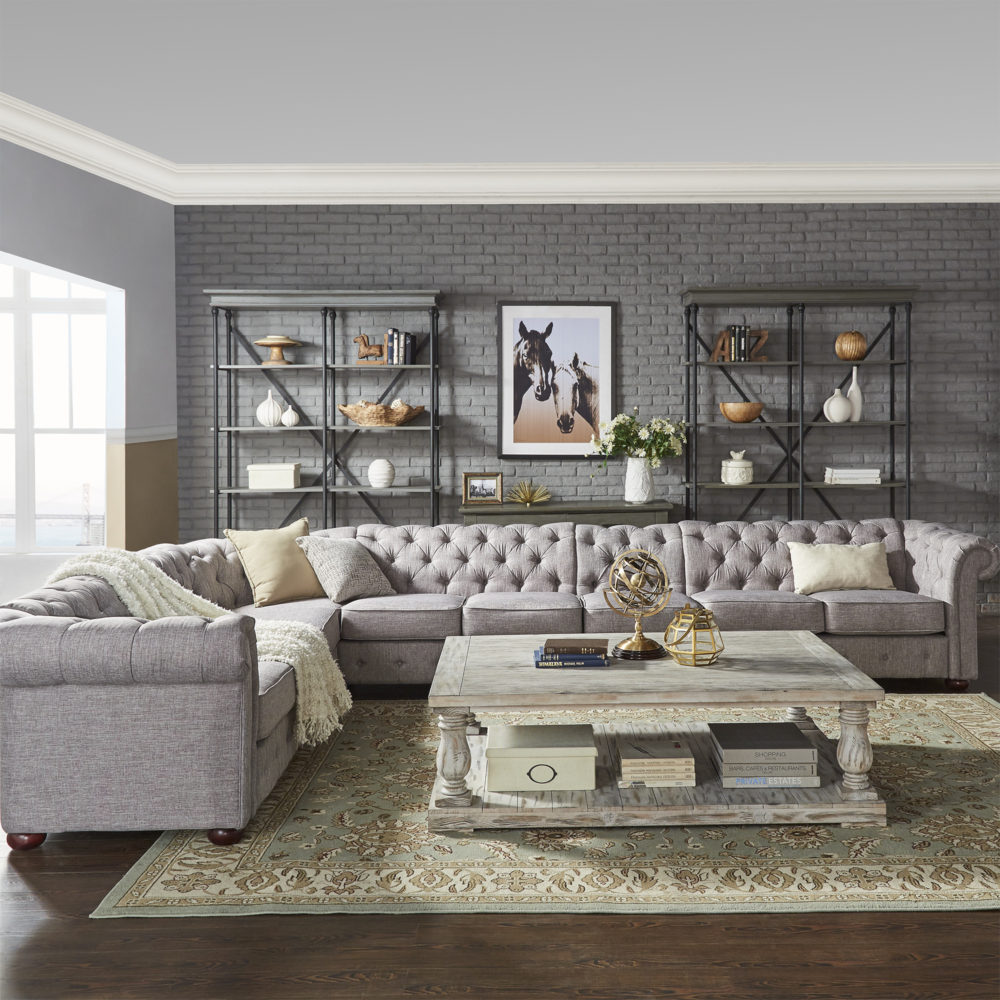 Another large living room exuding Old World grandeur. This living room setup features an L-shaped chesterfield sectional sofa with grey linen upholstery and soft button tufting. With a neutral and light color scheme, the rectangular wood coffee table has a distressed white finish, while the wood sofa table and wood and metal bookcases boast a light grey finish.