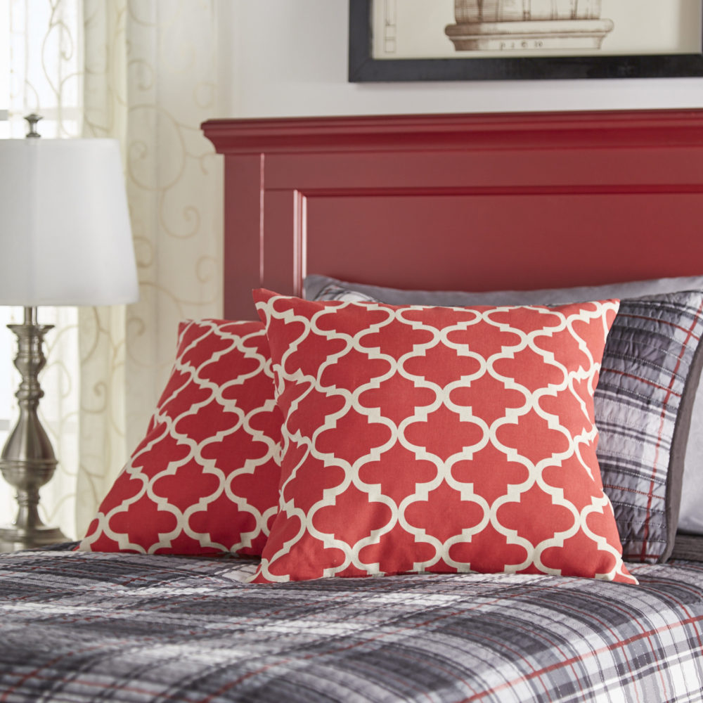 A kid's bed with a bright red wood panel headboard. The bed is dressed with a grey, black, and red plaid print blanket and pillows. There are also bright red accent pillows with a Moroccan print.