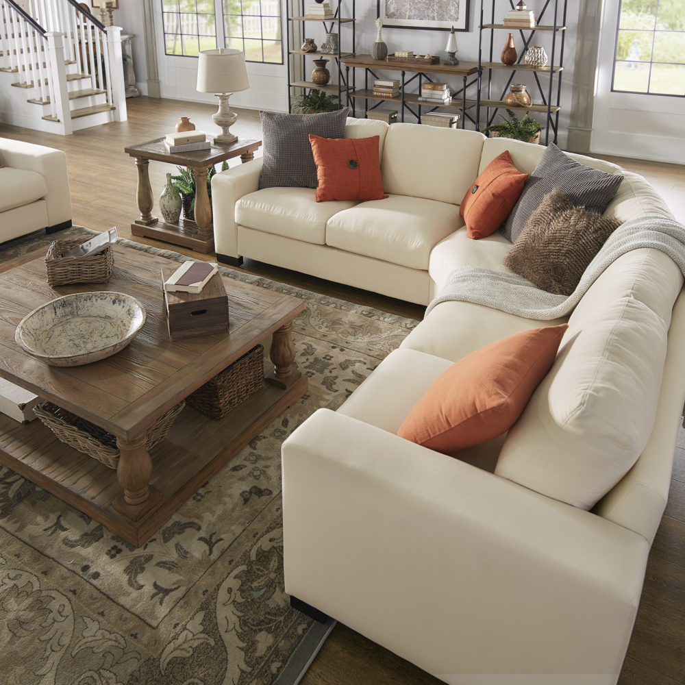 Another view of the white fabric upholstered sectional sofa. There are a variety of throw pillows on the sofa, all of various shades of orange and brown. These tones pair well with the natural wood grain finish of the coffee table, end table, and bookcases creating a warm atmosphere. 