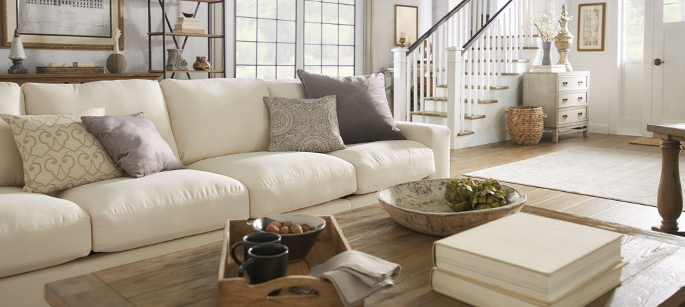 This is a closeup view of our classic-styled space. There is a plush, white fabric sofa with neutral grey throw pillows decorating it. In front of the sofa is a brown wood rectangular coffee table with some rustic pieces of décor. These neutral pieces blend well with the white walls and brown hardwood floors.