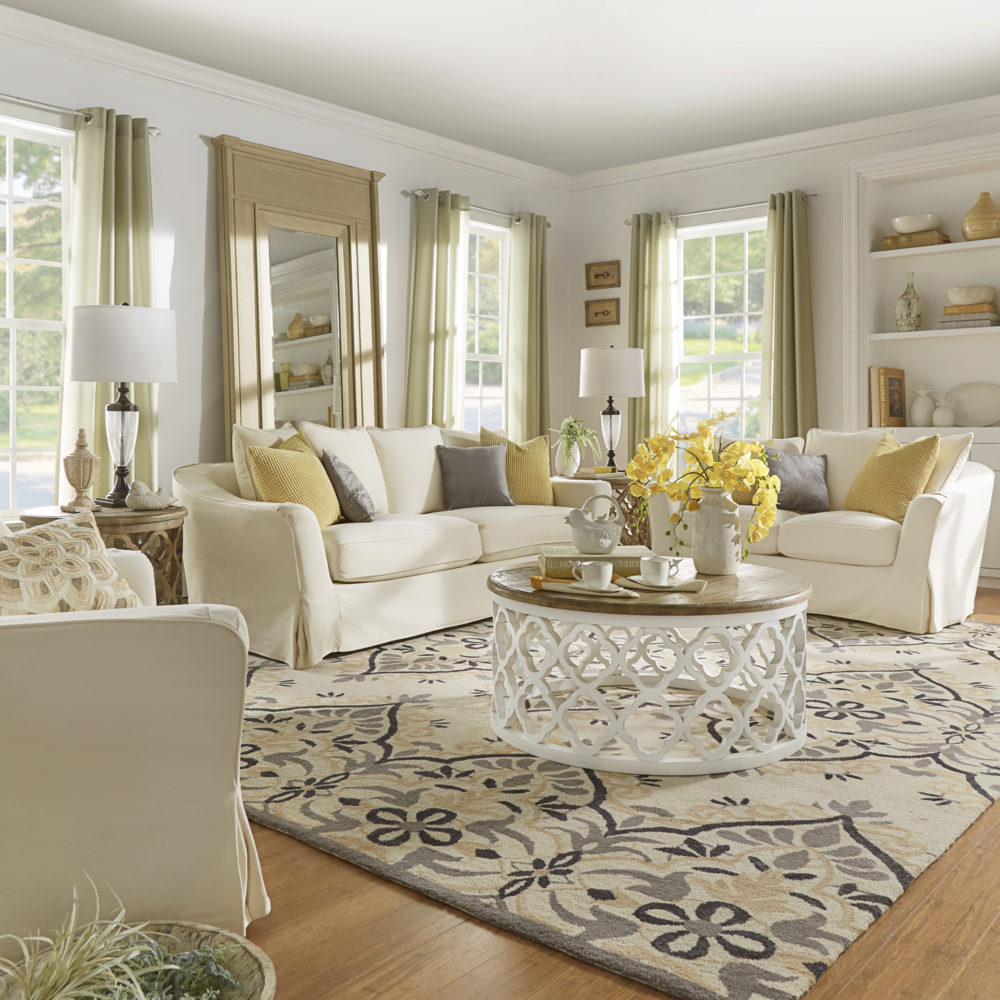 A light and fresh living room. There are white cotton slipcovered sofa, loveseat, and accent chair around a white and oak round coffee table. The room is decorated with yellow, grey, white, and green décor that is effortlessly timeless.