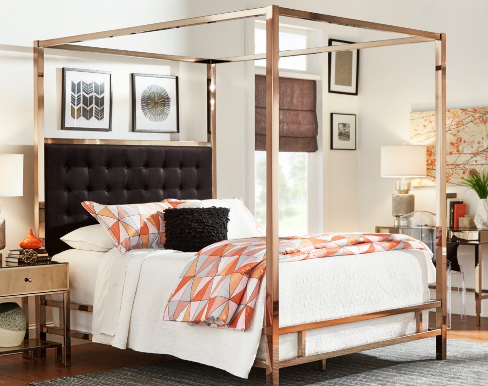 Another bedroom displaying how to mix textures. The bedroom features a canopy bed with a champagne gold metal frame and black button tufted upholstered headboard. The bed is dressed with mostly white bedding, but pops of color come from multicolored blankets, pillows, and a black accent pillow. There is a mirrored 1-drawer nightstand decorated with various pieces of décor. Against the adjacent wall is a matching mirrored 1-drawer writing desk with a clear acrylic chair. 