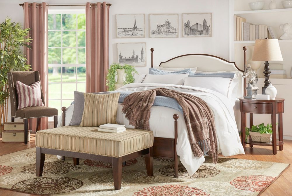 A classical-themed bedroom. The poster bed has a wood frame with a cream fabric upholstered headboard. The bed is dressed with blue and white bedding, as well as a plaid brown throw blanket. There is a brown wood nightstand with various pieces of décor on it. At the foot of the bed is an ottoman with a wood base and green striped fabric upholstery. Off to the side is a brown fabric upholstered wingback parsons chair with a silver nailhead trim. The room also has various greenery throughout.