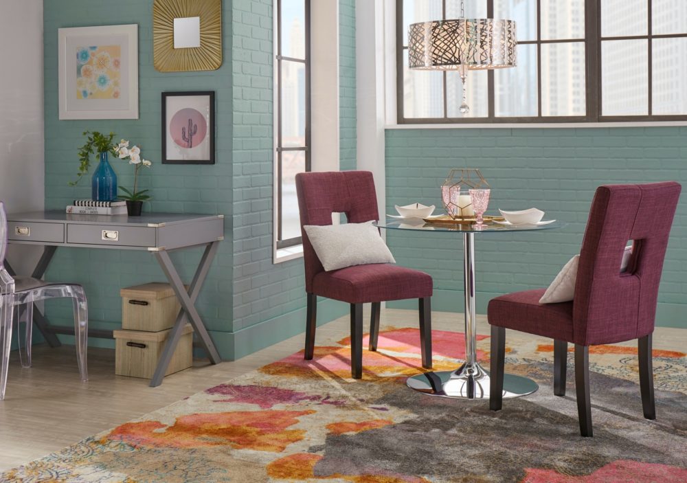 A corner of a living space that shows how to mix textures. On the left side is a grey finish campaign writing desk and a clear acrylic chair. On the right side is a round glass top dining table with a chrome metal base. Paired with the dining table are two keyhole dining chairs upholstered in tawny port red linen.