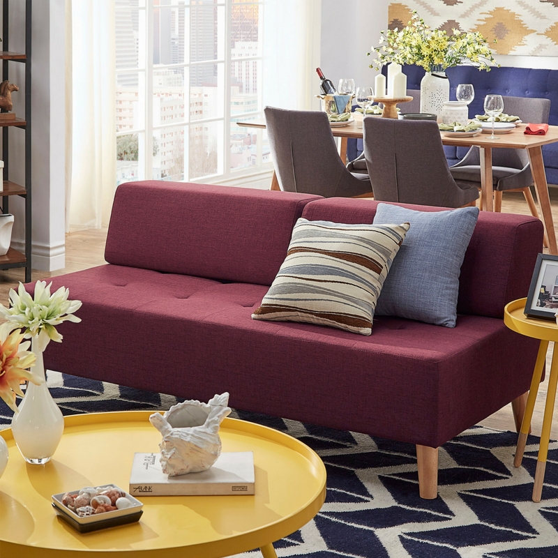 This image is another living room. In the center is a tawny port red armless sofa, minimally decorated with two accent pillows. The accent pillows are blue and brown, contrasting against the red fabric. In the background is a Scandinavian-inspired dining set. In the foreground is a bright yellow tray top coffee table.