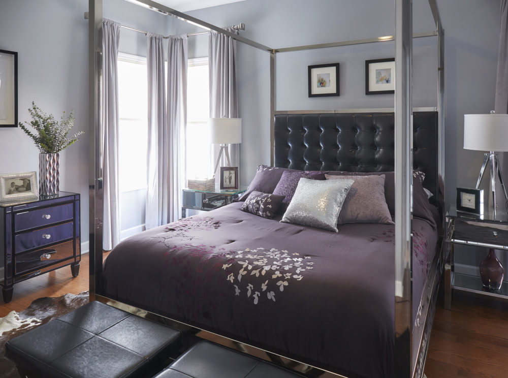This is one of the bedrooms part of our Illinois home makeover. The centerpiece is a stunning canopy bed with a chrome metal frame and a black bonded leather button tufted headboard. This bed is decorated with bedding of various purple shades, primarily deep purple to complement the black headboard. At the foot of the bed are two stools, both of which are upholstered in black faux leather. There are three mirrored nightstands in the room, two of which are by the bed while the third is a 3-drawer piece off to the side.