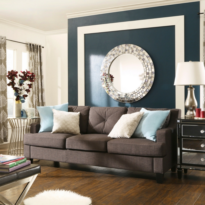 This image shows another living room. The dark grey linen tufted sloped track arm sofa sits in the center of the room. There are four accent pillows on the sofa, two light blue ones, and two grey and white ones, offering a delicate contrast against the dark grey linen of the sofa. On the back wall is a large, round wall mirror. Each side of the sofa has a mirrored and chrome finished end table, elevating the look of the room.