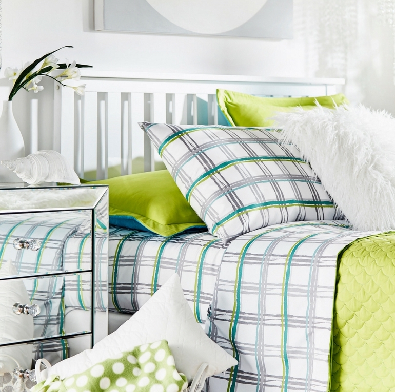 This image features the first of our kids and teen bedroom ideas. It shows a white bed with a slatted headboard. The bed is dressed with green, blue, white, and grey bedding. The sheets and a pillow have a plaid-style print with these colors. Other pillows are solid green, blue, and white. Next to the bed is a mirrored 3-drawer nightstand. 