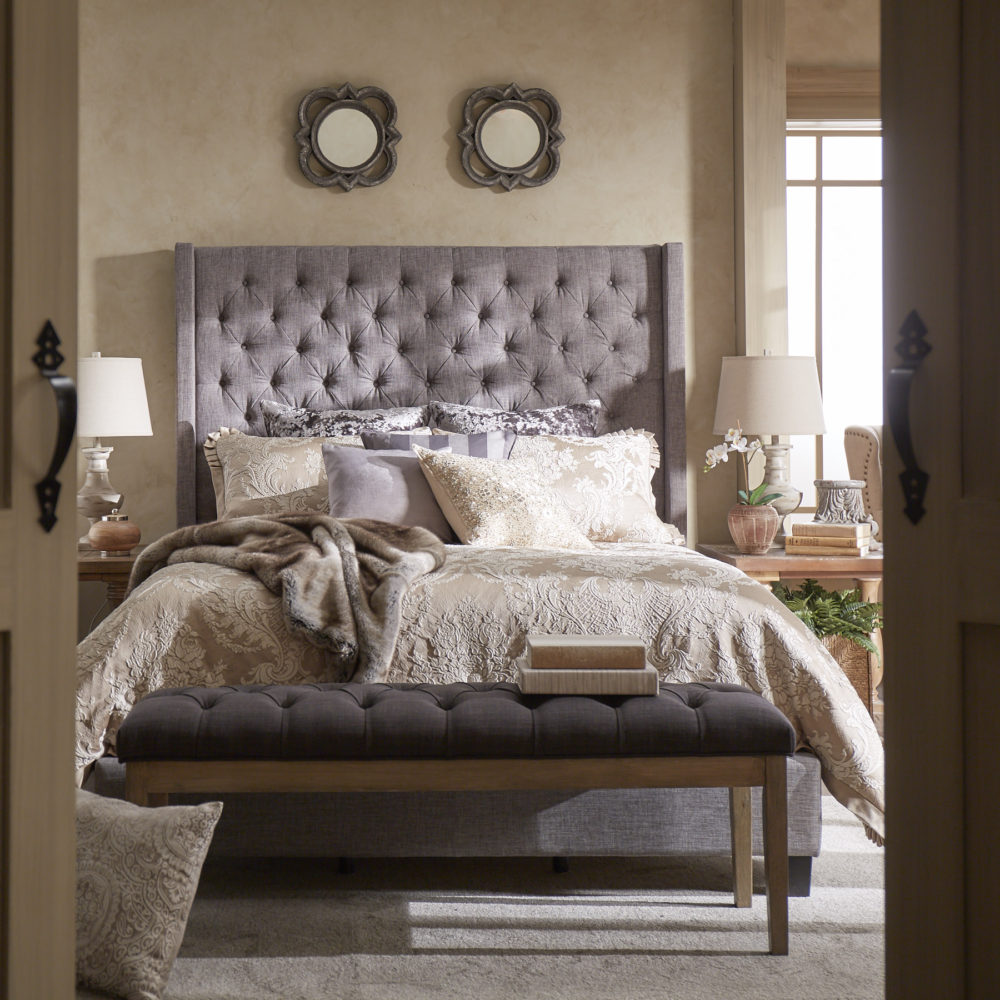 This is another look to show how to style an upholstered headboard. This bed from our iNSPIRE Q Artisan collection is upholstered in grey linen and features a wingback, button-tufted headboard. This bed features more elaborate bedding. It uses beige and grey colors, with paisley prints on both the comforter and the pillows.