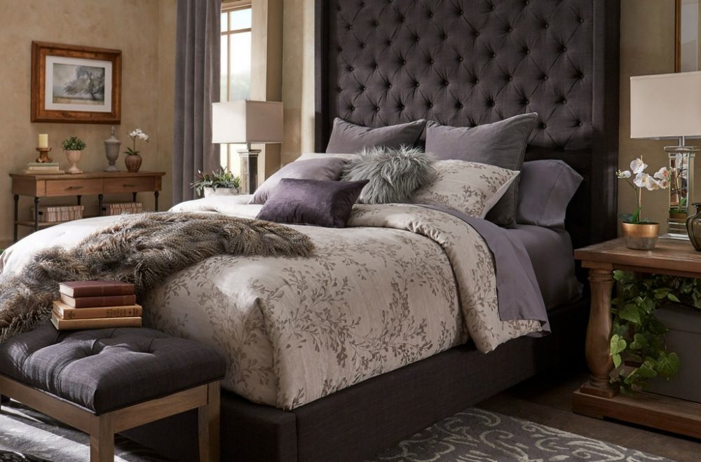 This image displays one of our beds from iNSPIRE Q Artisan. The bed is upholstered in dark grey linen and has an extra tall, wingback headboard with deep button tufting. The bed features floral-themed bedding, with plenty of pillows in various shades of grey, purple, and white. The sheets are also purple, which complements the dark grey linen upholstery.