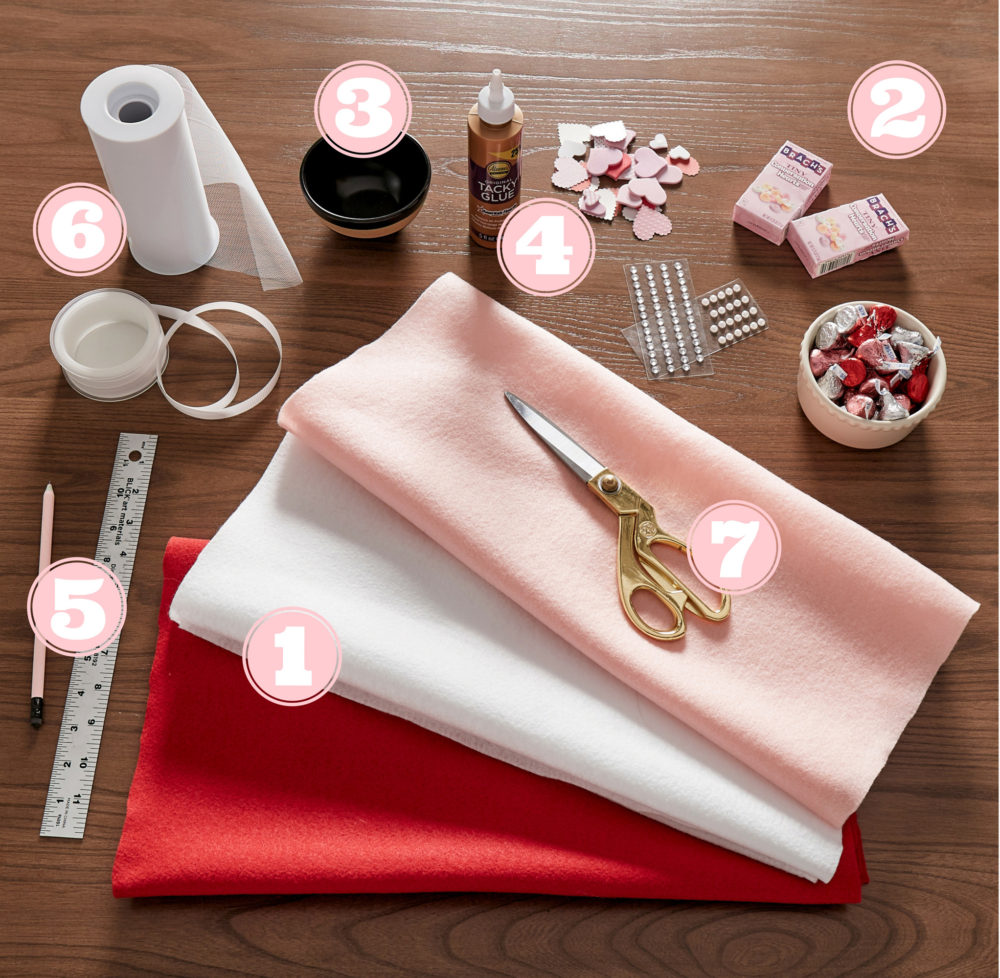 This image shows all the items needed to create a DIY Valentine's Day table. There is fabric, a ruler, a pencil, ribbon, glue, tiny hearts, and scissors, among others. All pieces are labeled with a number to correspond with the list.