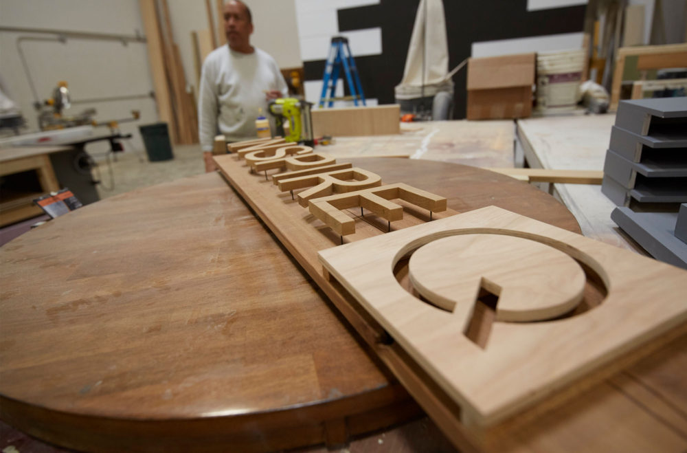 This image depicts a custom-made wooden sign of the iNSPIRE Q logo. The sign is not yet hung up, but sits on a table with one of our team members in the background.