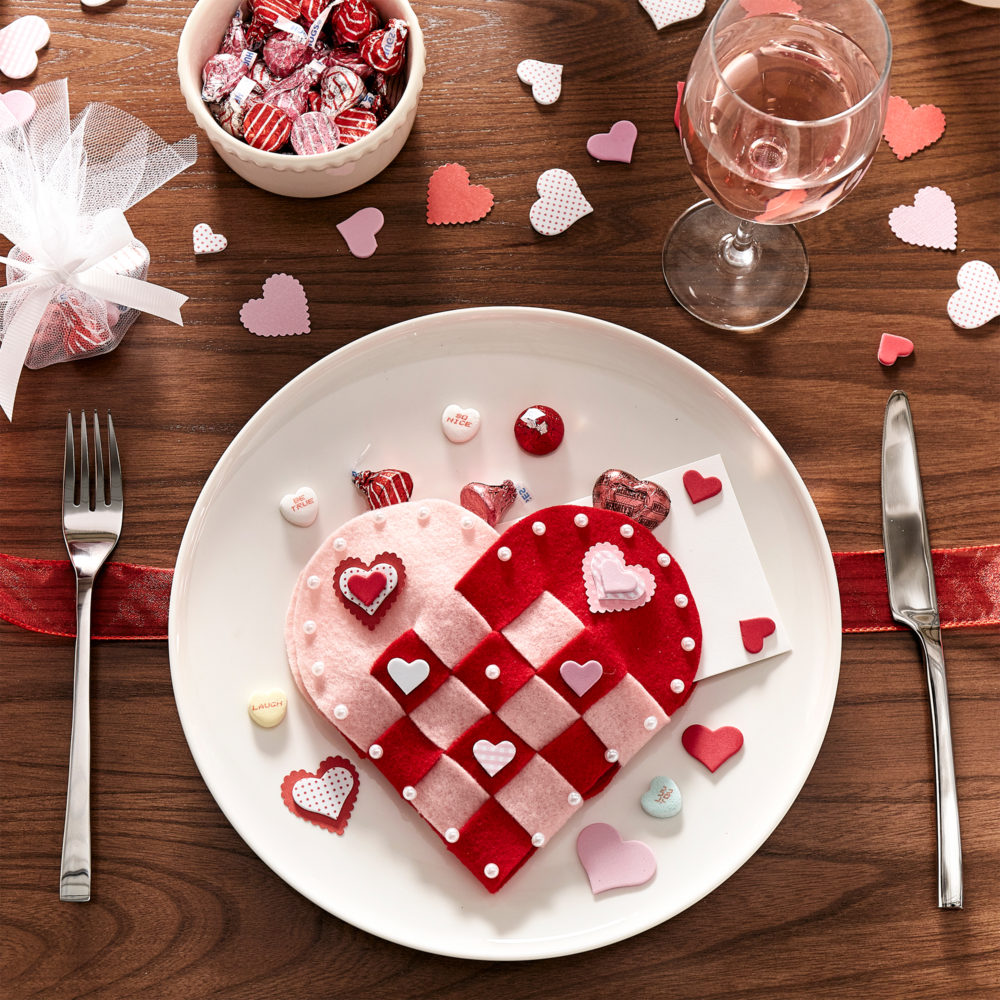This image shows the woven fabric heart on a place setting for our Valentine's Day table. There are hershey kisses, decorative confetti hearts, candy hearts, all on top of a white plate.