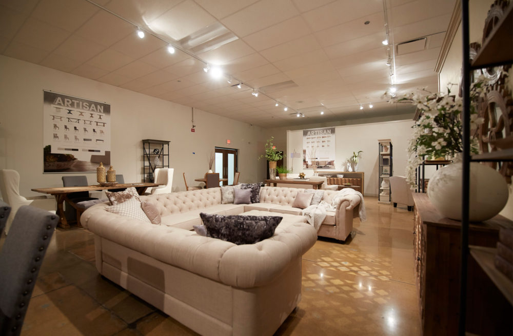 This image is another angle of our iQ Artisan section with a beige linen U-shaped sectional sofa as the centerpiece. In the background are two dining sets from the iQ Artisan collection, as well as some bookcases, console tables, and accent chairs. Hanging on the wall in the background is a poster listing some key pieces of the Artisan Collection.