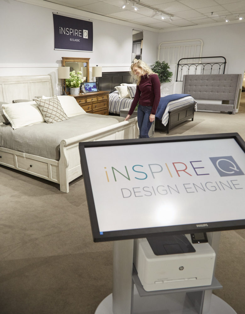 This photo shows another view of our Design Engine at Darvin Furniture. We see in the foreground the kiosk, while in the background is one of our team members admiring the wood sleigh and panel beds.