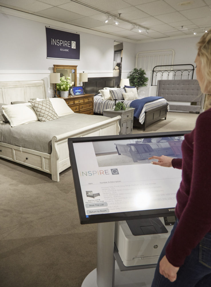 Here we see one of our team members using the Design Engine at Darvin Furniture. She is using the touch screen of the kiosk to browse the different options for the antique finish wood sleigh beds displayed behind the kiosk.