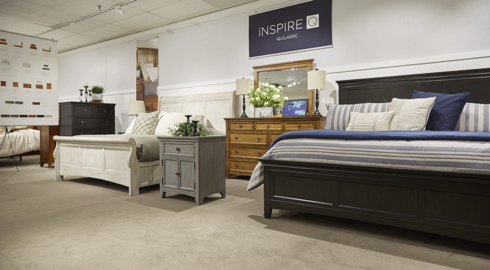 This photo displays a wide shot of the iNSPIRE Q Classic bedroom collection. It includes an antique black wood panel bed, an antique white wood sleigh bed, an oak finished dresser with mirror, and an antique grey wood nightstand.