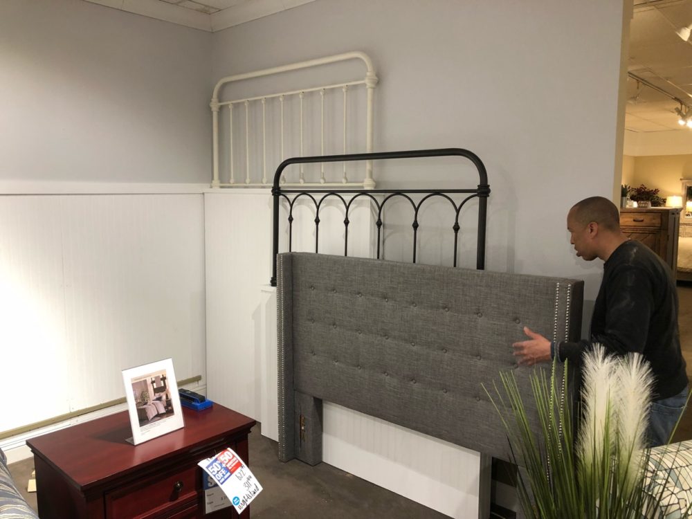This photo shows our team member setting up the wall display of headboards. There are three headboards pictured: the Antique White Graceful Lines Victorian Metal Headboard, Black Casted Knot Metal Headboard, and Nailhead Wingback Tufted Grey Linen Upholstered Headboard.